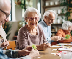 A group of elderly people are sitting around a table, smiling and enjoying their time together. They are engaged in a craft activity, working on paper flowers. The table is filled with various items, including cups, bowls, and potted plants, creating a cozy and welcoming atmosphere. AI generated content