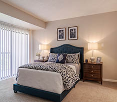 A bedroom with a bed, nightstand, and dresser. The bed has a blue comforter and pillows. A picture frame is on the headboard. AI generated content