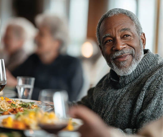 A man is sitting at a table with a plate of food in front of him. He is smiling and appears to be enjoying his meal. There are other people in the room, some of whom are also seated at the table. There are wine glasses and cups on the table, suggesting that the people are having a meal together. AI generated content